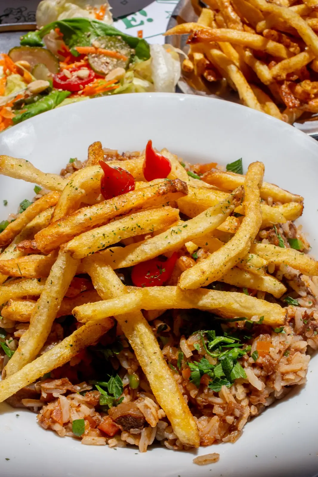 Prato de arroz temperado coberto de batatas fritas, com salada ao fundo