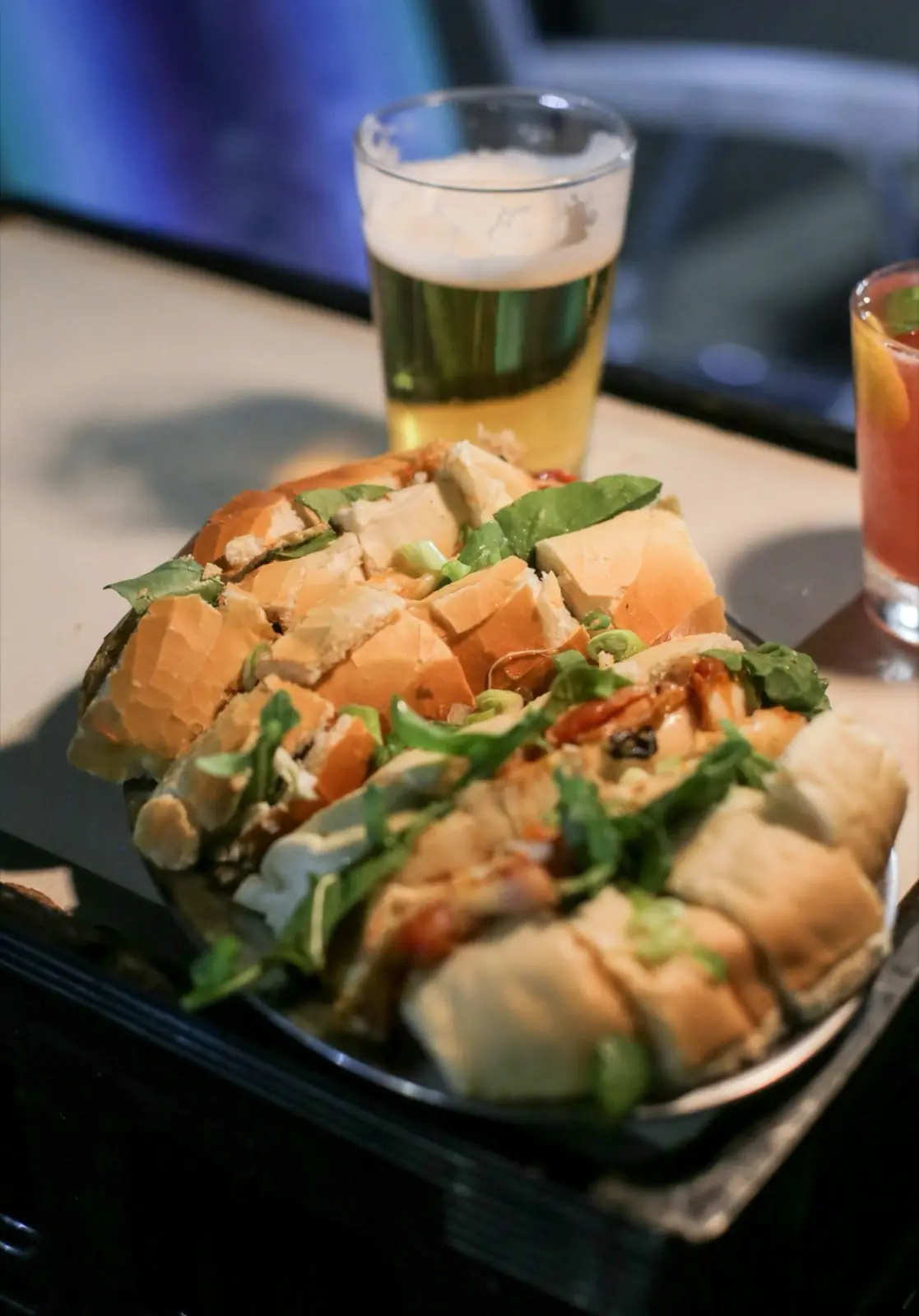 Pão recheado e fatiado, servido com um chope e um drink ao fundo