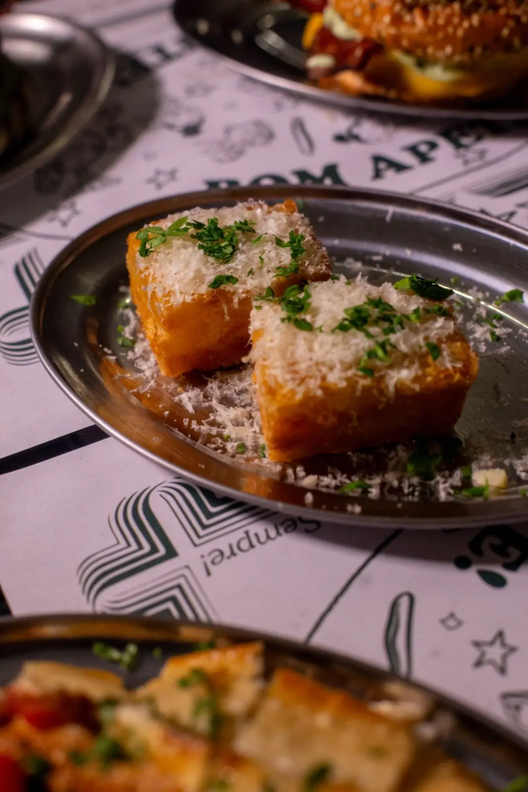 Fatias de pão dourado cobertas de queijo ralado e salsinha, num prato de metal